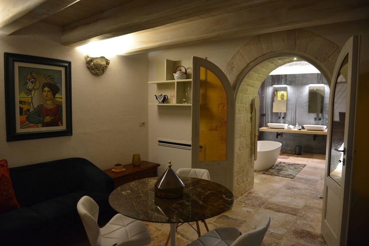 Una sala de estar moderna con una mesa redonda de mármol, un sofá azul y un cuadro en la pared. Una puerta abierta da a un baño con un lavabo grande y un espejo.