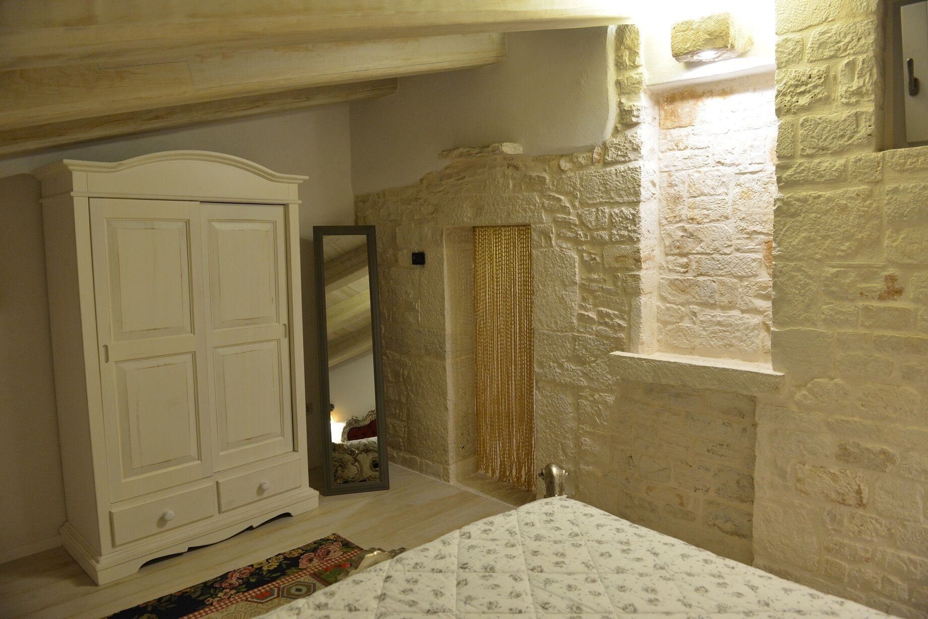 Une chambre avec une armoire blanche, un lit recouvert d’une couvre-lit à motifs, un miroir et un mur en pierre.