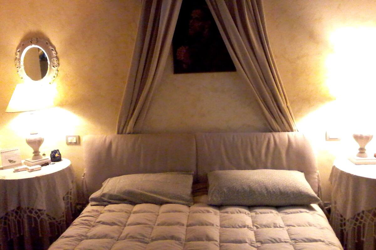Un lit double à baldaquin, deux tables de chevet avec des lampes, un miroir rond et une petite horloge au mur.