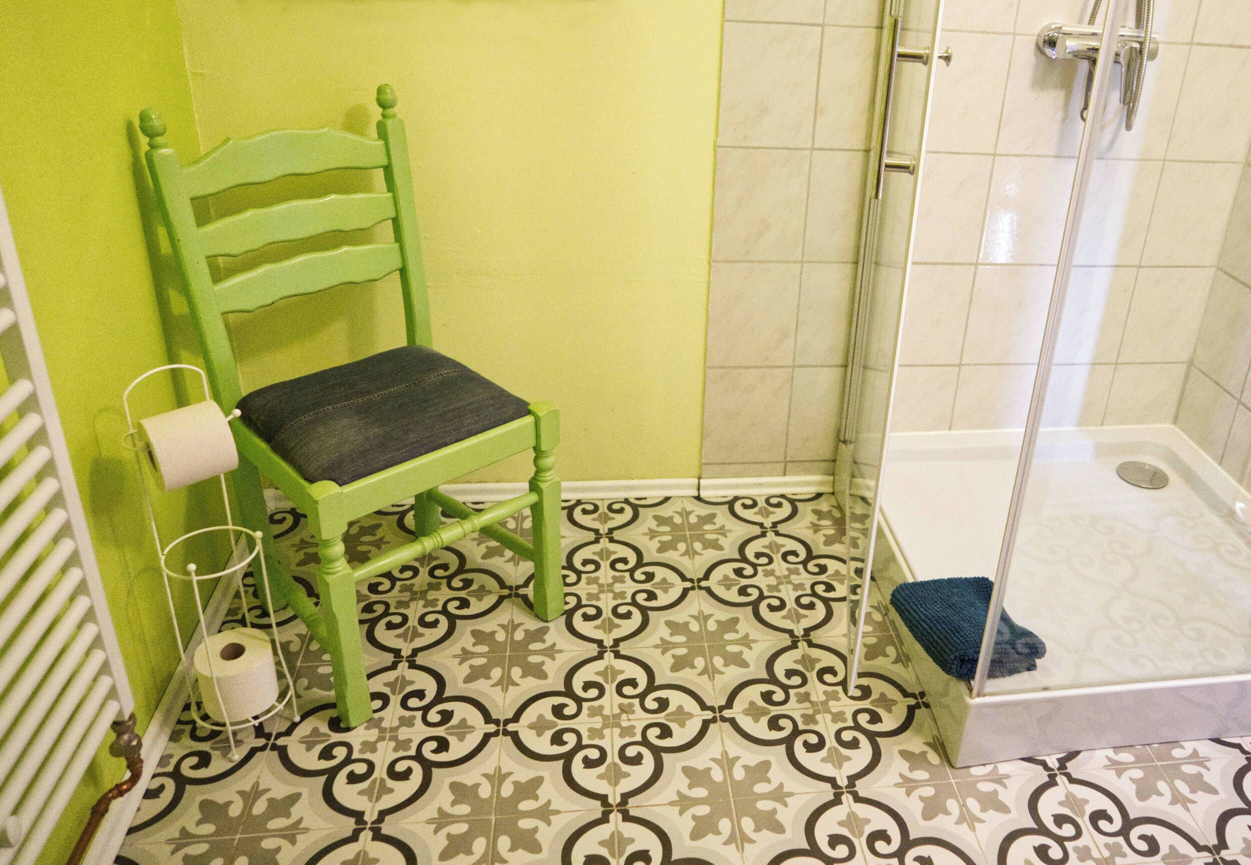 A bathroom with a green chair, a toilet, a shower, and patterned floor tiles.