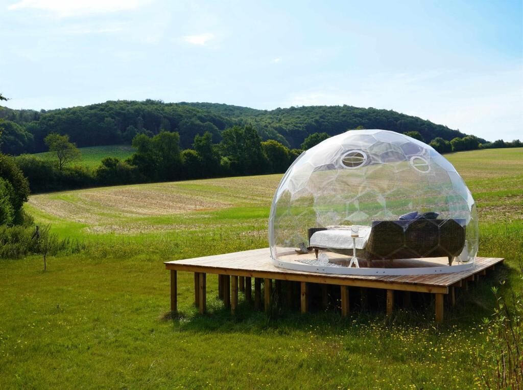 A transparent dome structure with a bed inside, set on a wooden platform in a grassy field with a forested hill in the background.