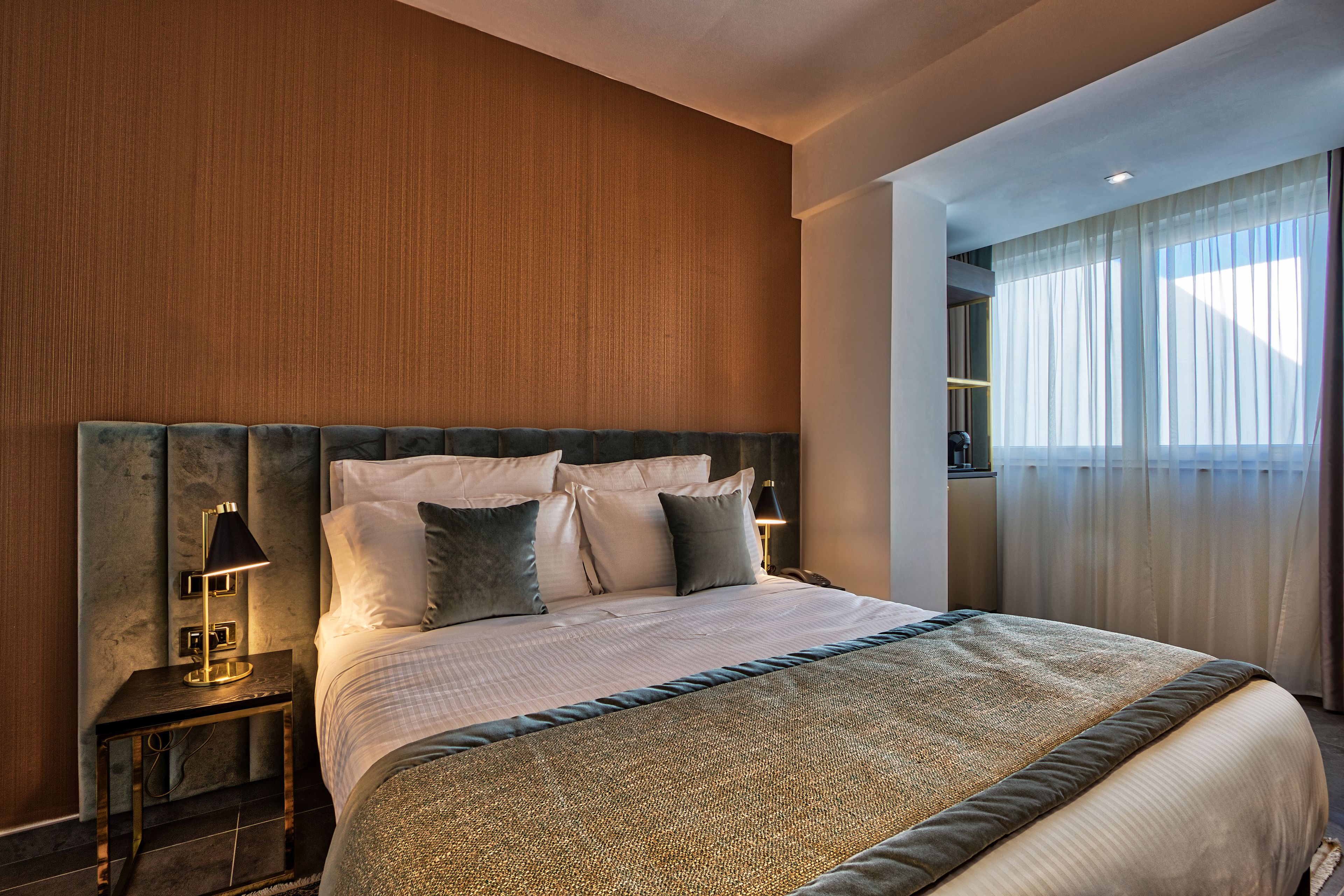 A modern hotel room with a large bed, a nightstand with a lamp, and a view of the outdoors through sheer curtains.