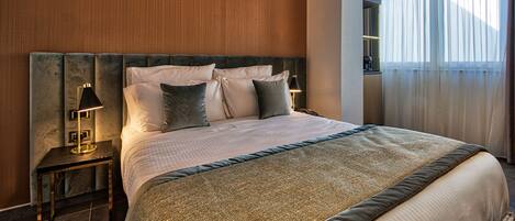 A modern hotel room with a large bed, a nightstand with a lamp, and a view of the outdoors through sheer curtains.