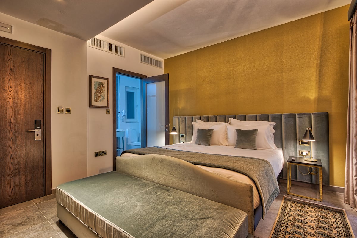 Ein modernes Hotelzimmer mit einem großen Bett, einem Nachttisch mit Lampe und Blick auf den Flur.