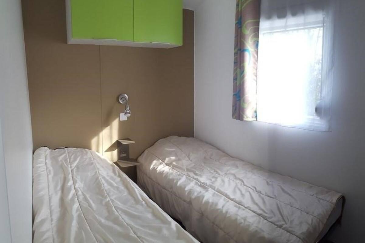 A small room with two single beds, a green cabinet, a window with a curtain, and a wall-mounted light.