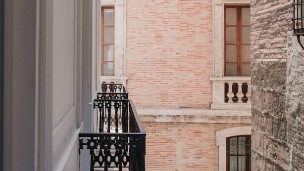 Un estrecho pasillo con pared de ladrillo y un balcón con barandilla negra ornamentada.