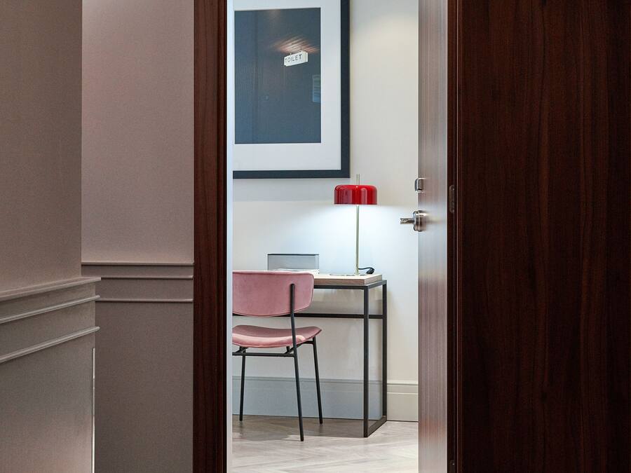 A hotel room entrance with a wooden door, a number '405' sign, a framed picture on the wall, a red lamp, and a pink chair.