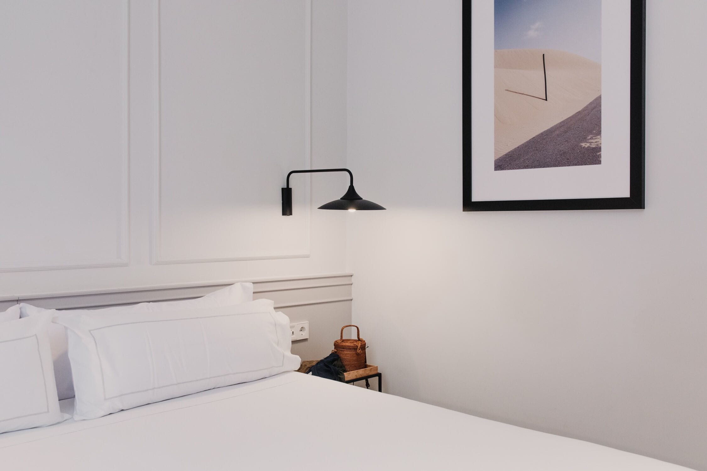 Camera da letto minimalista con un letto, una lampada da comodino, un quadro incorniciato e un piccolo tavolo in legno con un cesto.