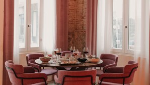 Una mesa de comedor dispuesta con sillas, copas de vino y vajilla, con una pared de ladrillo y ventanas al fondo.