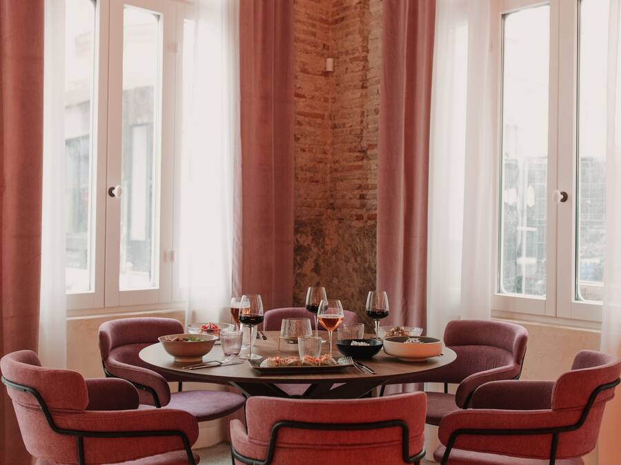 Una mesa de comedor dispuesta con sillas, copas de vino y vajilla, con una pared de ladrillo y ventanas al fondo.