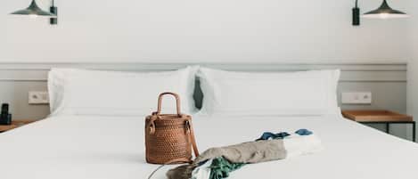 A neatly made bed with white linens, a brown woven bag, and a folded towel on a white wall with two black wall-mounted lamps.