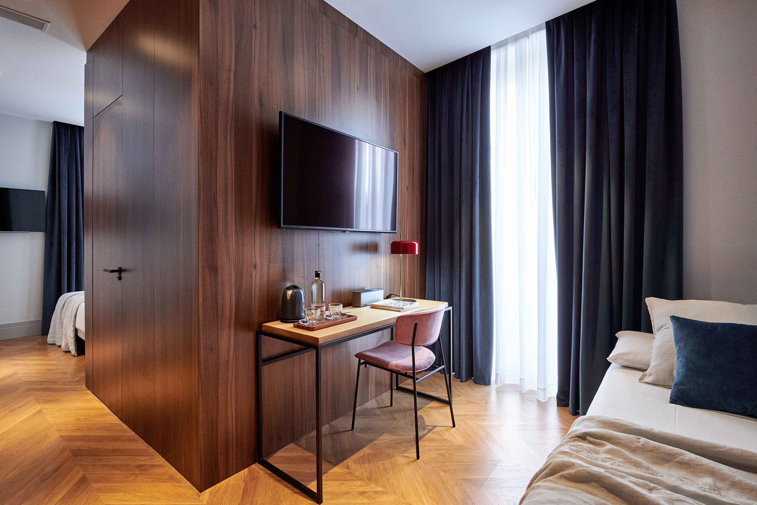 A modern hotel room with a wooden accent wall, a flat-screen TV, a desk with a chair, a bed with pillows, and a large window with curtains.