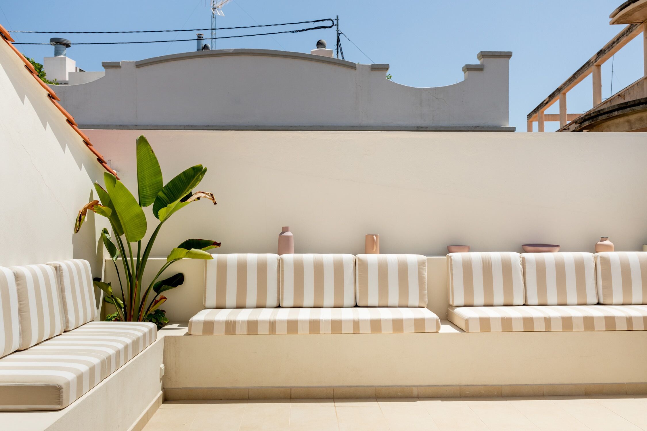 Un área de estar moderna al aire libre con un sofá a rayas blanco y beige, una planta grande y jarrones decorativos.