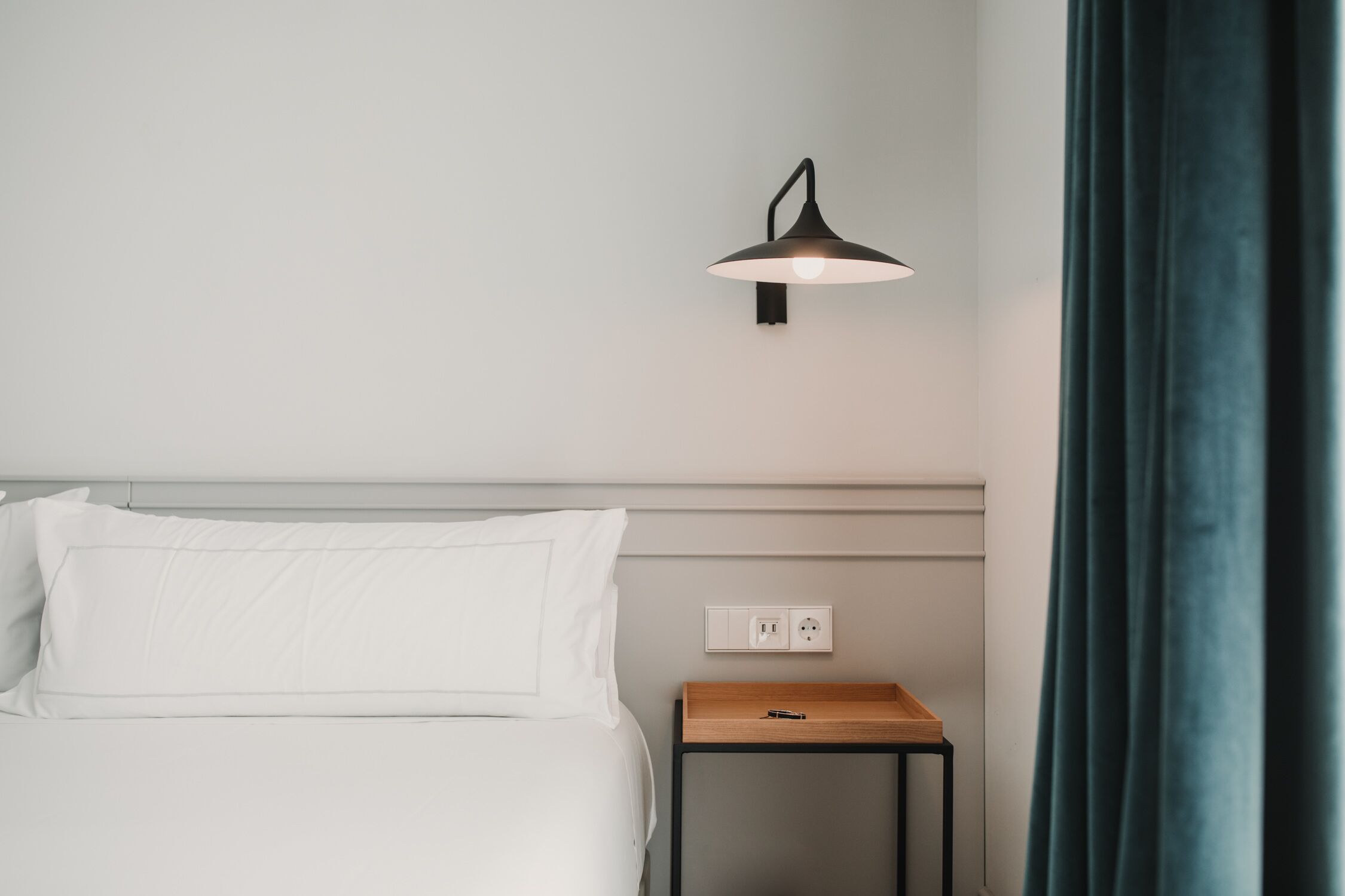 A neatly made bed with white linens, a bedside table with a lamp, and a power outlet on the wall.