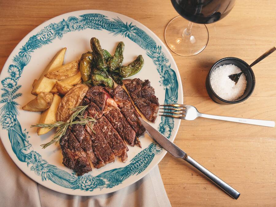 Prato com bife, batatas assadas e pimentões grelhados em cima de uma mesa, com um copo de vinho e saleiros.