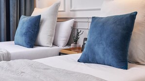 Habitación de hotel con dos camas, cada una con almohada blanca y azul, una mesita de noche con lámpara y una ventana con cortinas.