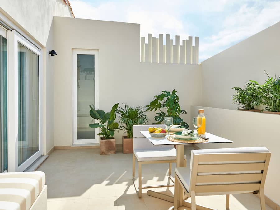 A modern outdoor dining area with a table set for two, surrounded by potted plants and a sliding glass door.