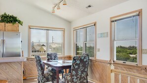 A dining area with a wooden table and floral chairs, large windows with views, and a kitchen with a refrigerator.