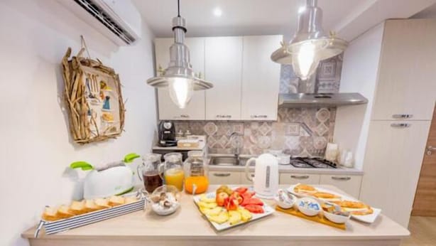 A modern kitchen with white cabinets, a central island with breakfast items, and pendant lighting. - B&B Dimora del Pescatore Cefalù (Cefalù)