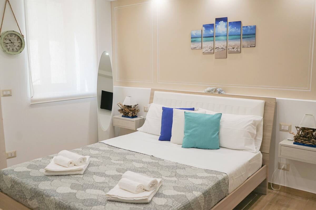 Un lit bien fait, recouvert d’une couvre-lit à motifs, une table de chevet avec une lampe et un miroir fixé au mur.