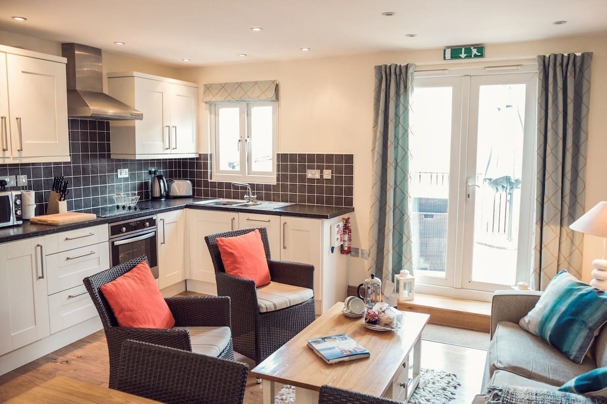 A modern kitchen with white cabinets, a black tiled backsplash, and a stainless steel range hood. There is a dining area with wicker chairs and a table, and a sofa with cushions.