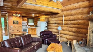 A log cabin interior with a leather sofa, two armchairs, a wooden coffee table, a kitchen area with a refrigerator and microwave, and a chandelier. - Infinite Views - 2BR/New Hot Tub/New Firepit/Grill/Sleeps 6 (Gatlinburg)