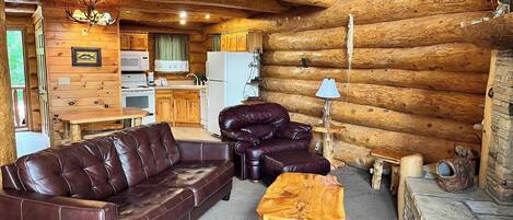 A log cabin interior with a leather sofa, two armchairs, a wooden coffee table, a kitchen area with a refrigerator and microwave, and a chandelier.