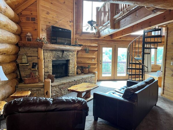 A log cabin interior with a stone fireplace, leather sofas, and a spiral staircase.