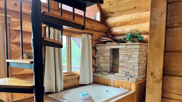 A wooden cabin interior with a stone fireplace, a wooden bathtub, and a window with curtains. - Infinite Views - 2BR/New Hot Tub/New Firepit/Grill/Sleeps 6 (Gatlinburg)