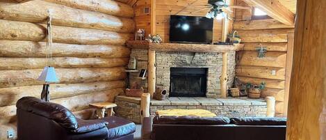 A log cabin interior with a stone fireplace, a television, and leather furniture.