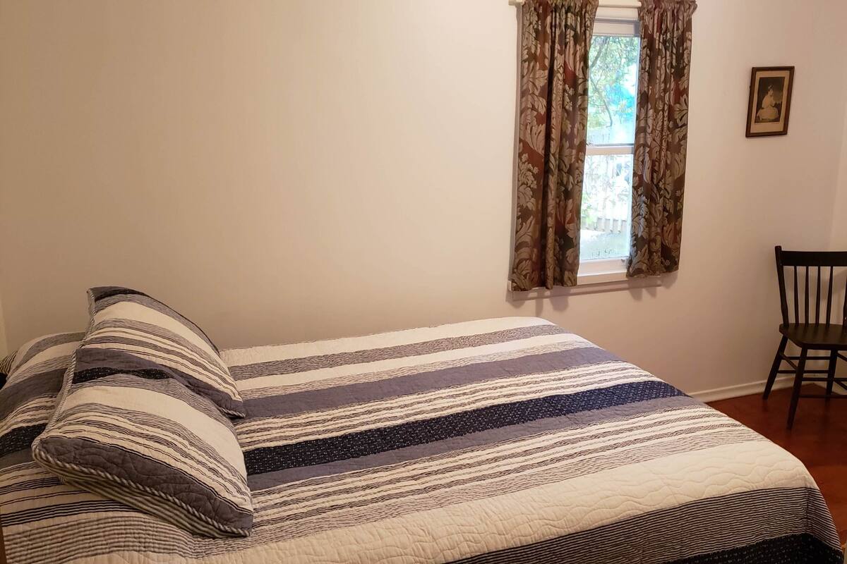 A bedroom with a bed, a window with curtains, a chair, and a framed picture on the wall.