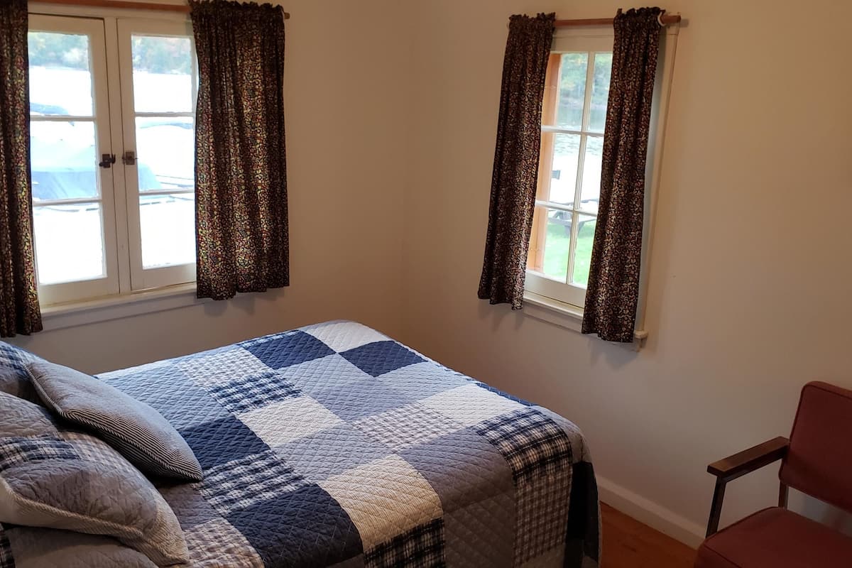 A bedroom with a bed, two windows with curtains, a chair, and a painting on the wall.