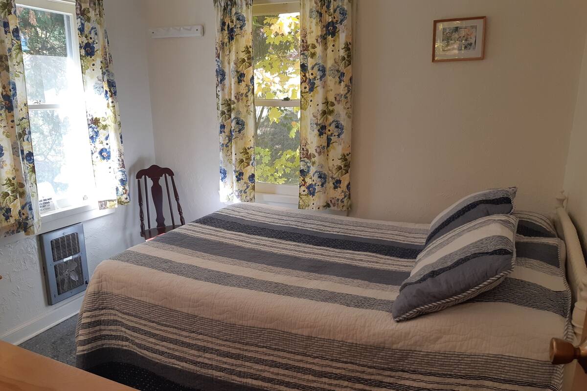 A bedroom with a bed, a wooden chair, a radiator, and a window with floral curtains.