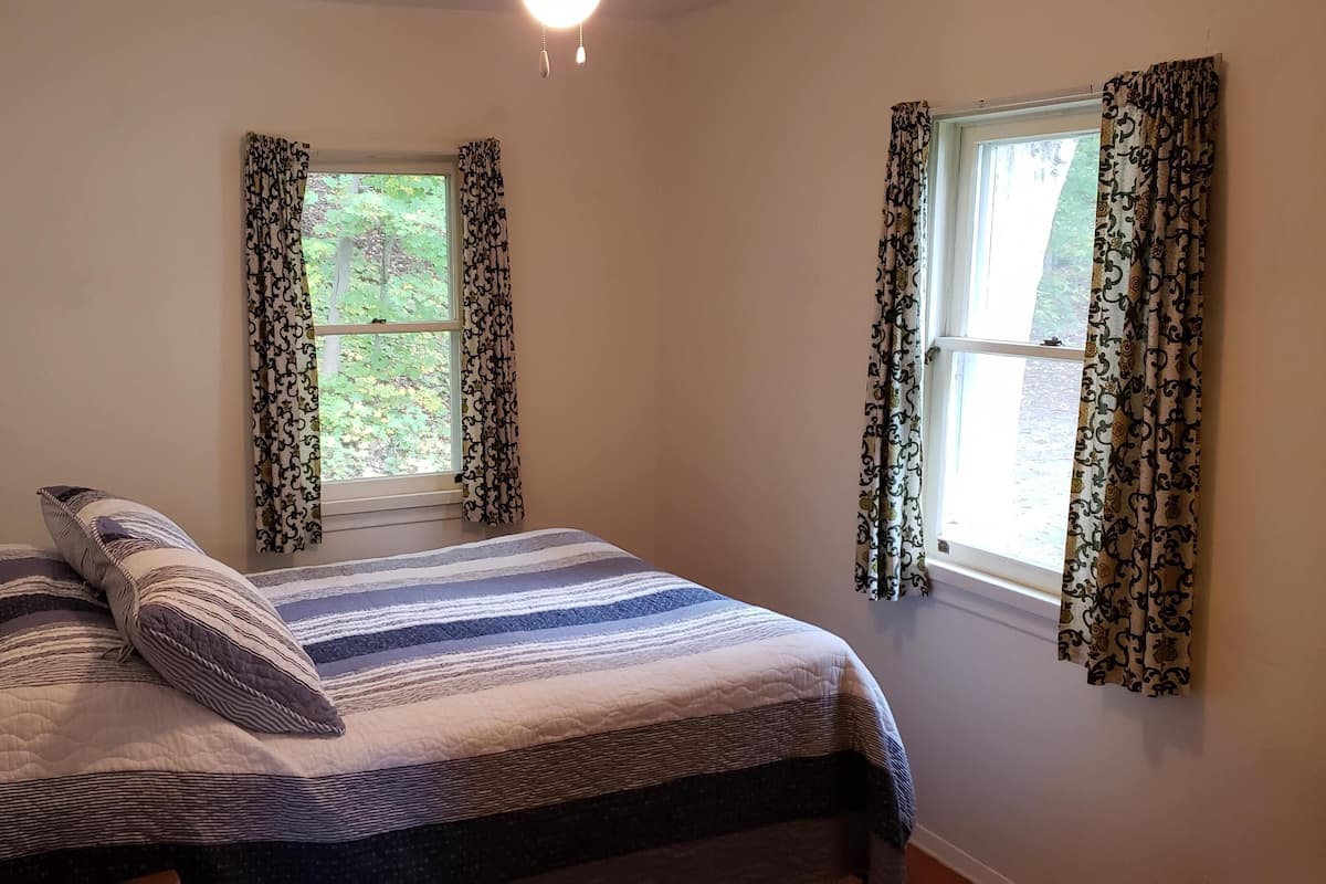 A bedroom with a bed, two windows with patterned curtains, a bedside lamp, and a view of greenery outside.