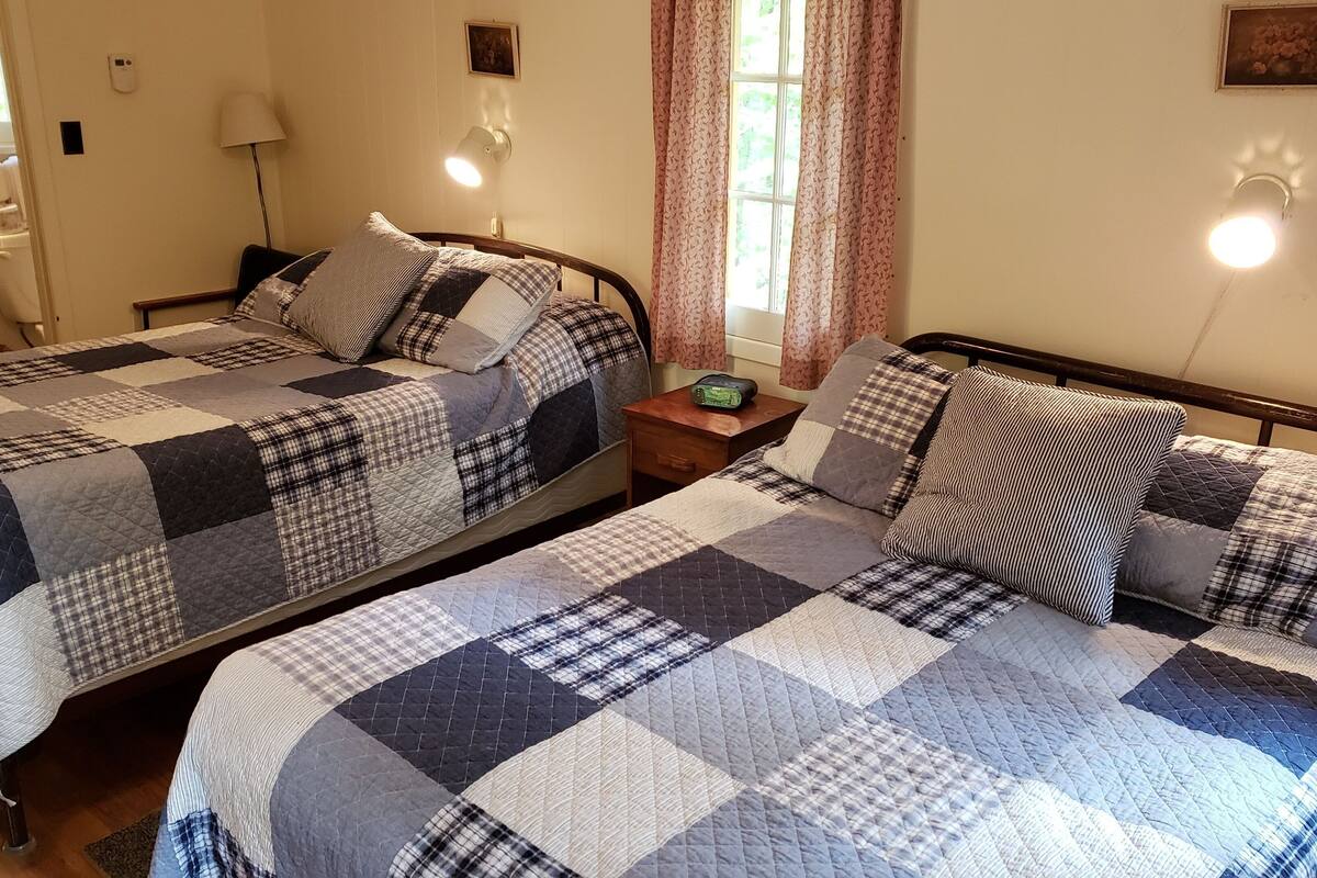 A room with two beds, each with a quilted bedspread and pillows. There is a nightstand with a radio between the beds.