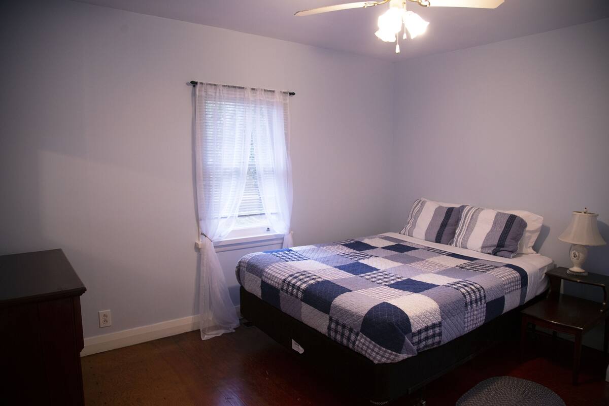 A bedroom with a bed, a nightstand, a lamp, a window with curtains, and a ceiling fan.