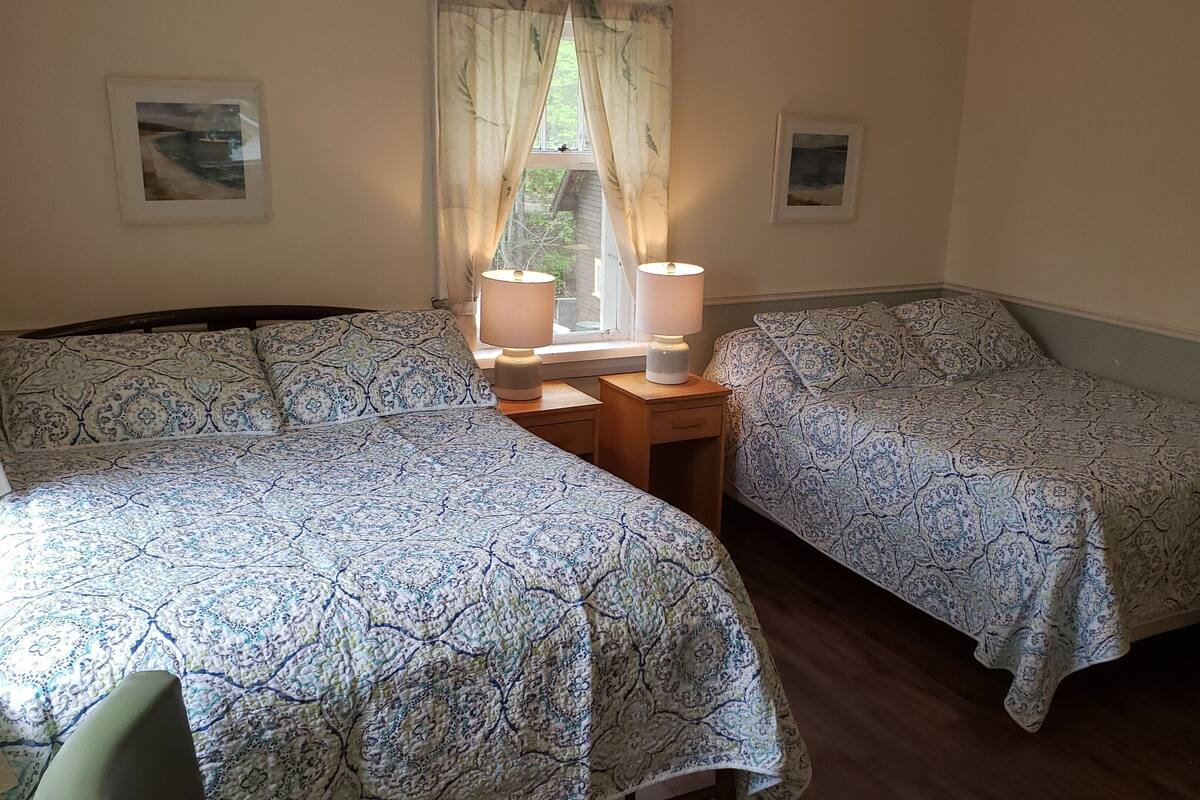 A room with two beds, each with patterned bedding, a window with curtains, and a nightstand with a lamp.