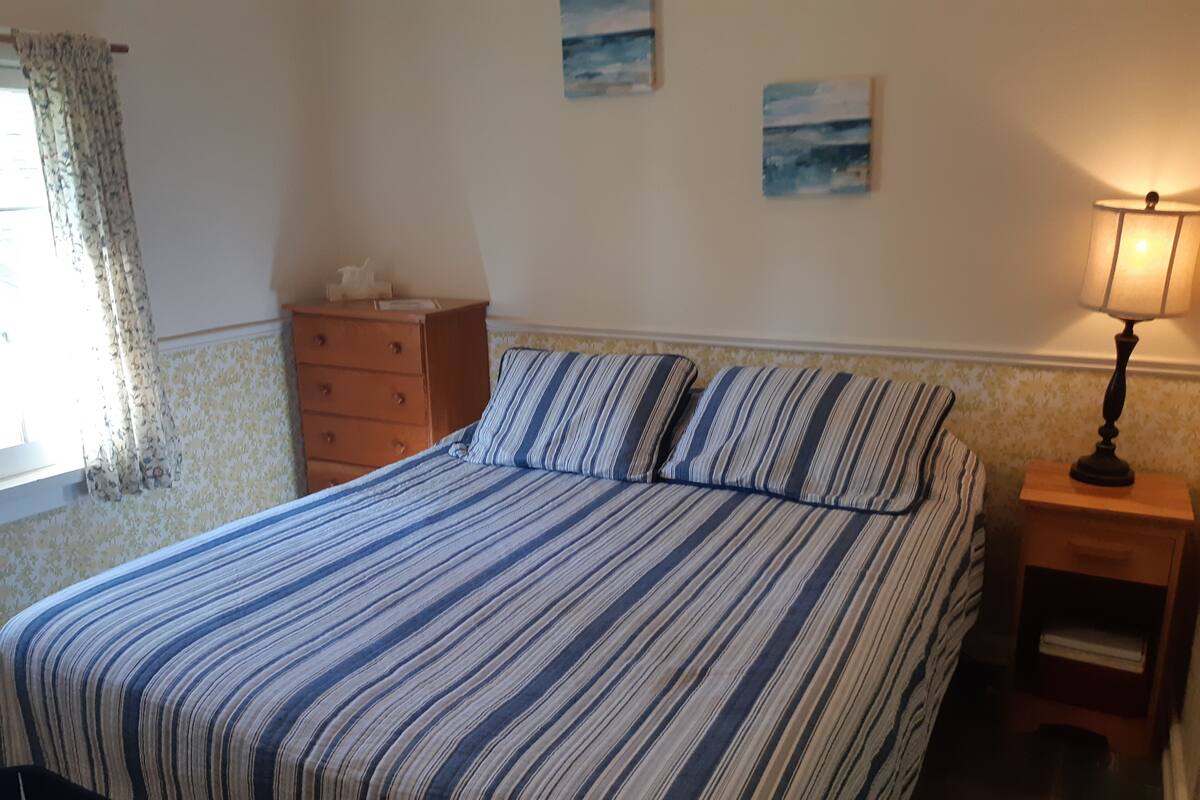 A bedroom with a striped bedspread, a wooden dresser, a nightstand with a lamp, and a window with curtains.
