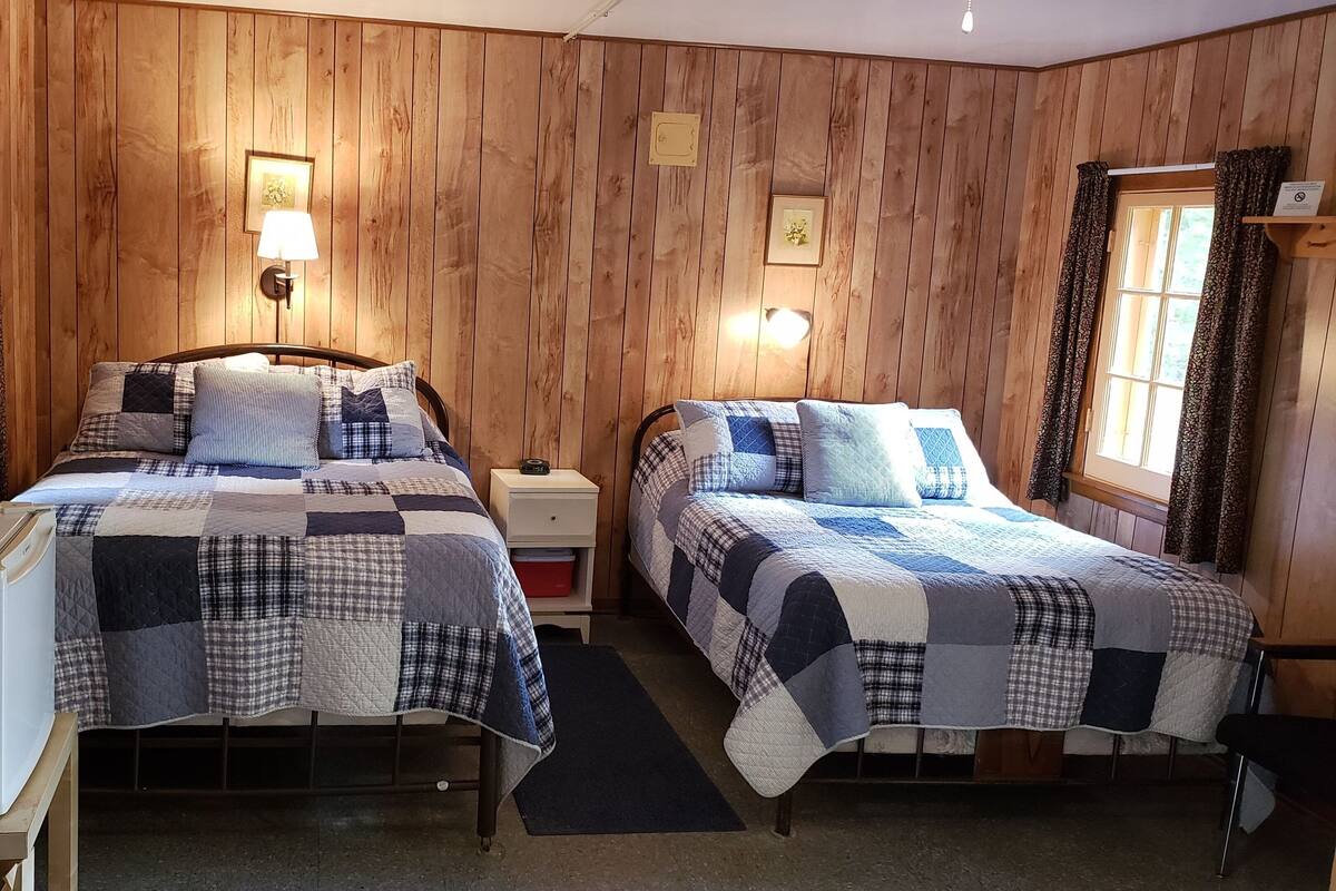 A room with two beds, a small table, a refrigerator, and wooden walls.