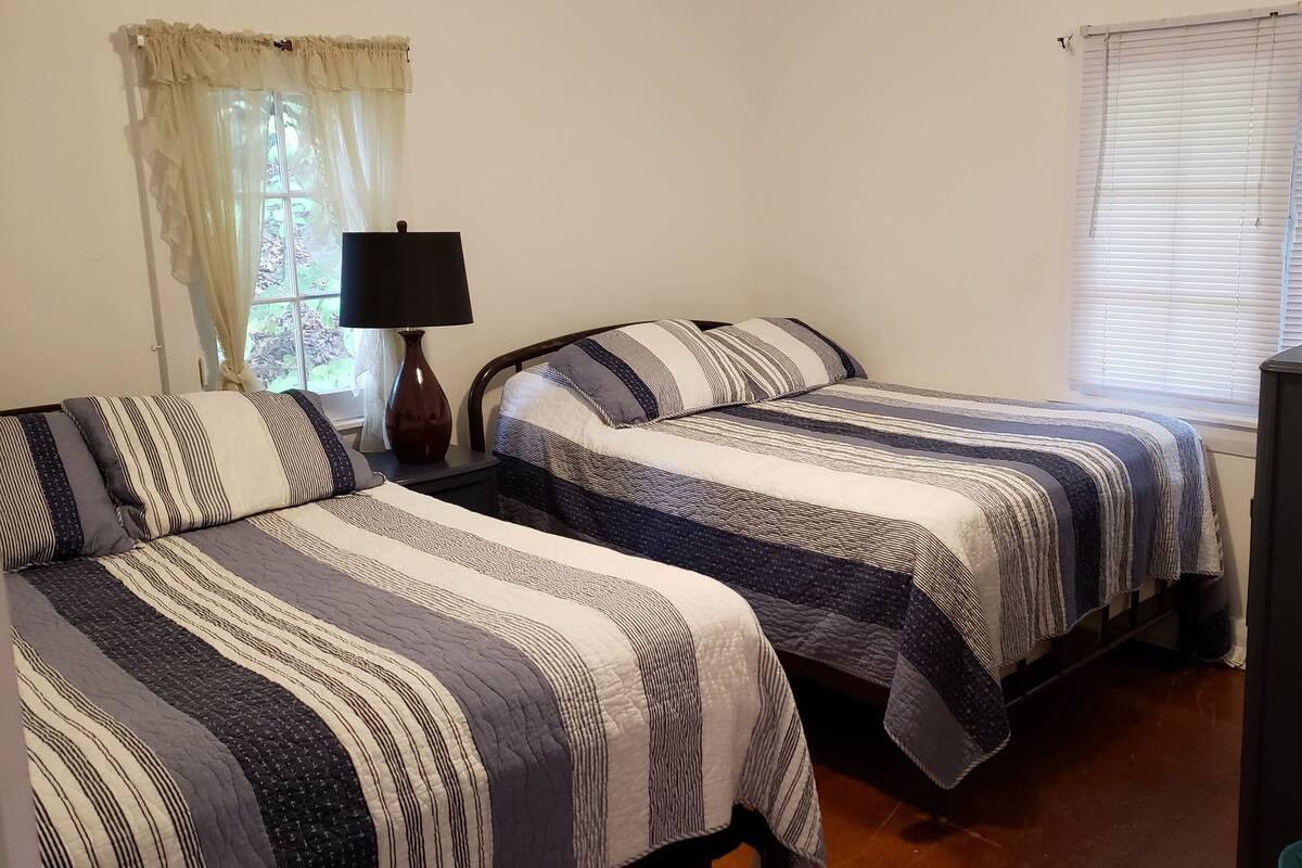 A bedroom with two beds, a nightstand with a lamp, a dresser, and a window with blinds.