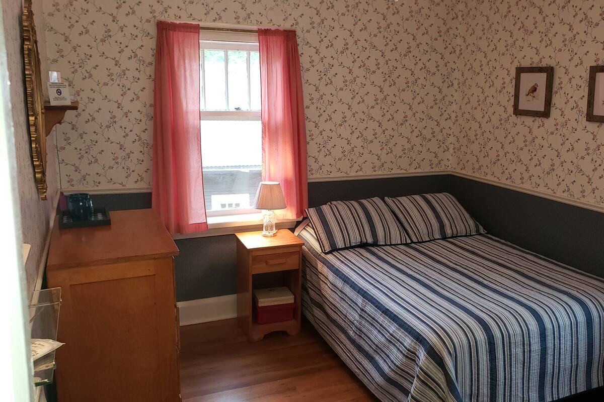 A small bedroom with a single bed, a nightstand, a window with curtains, and a wall with floral wallpaper.