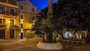 Una plaza urbana iluminada con luz cálida, con una palmera en el centro y edificios circundantes con balcones.