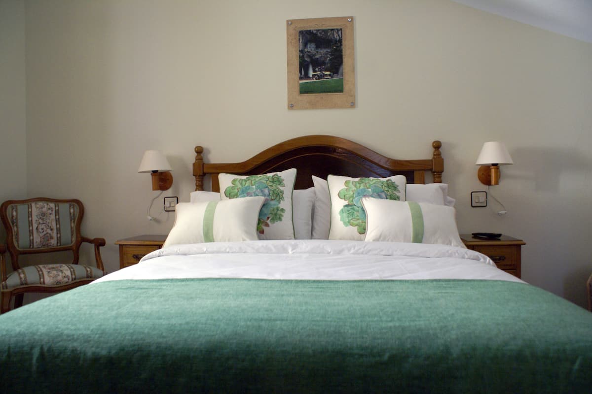 A neatly made bed with a green bedspread and floral pillows, flanked by two bedside tables with lamps, a chair, and a framed picture on the wall.