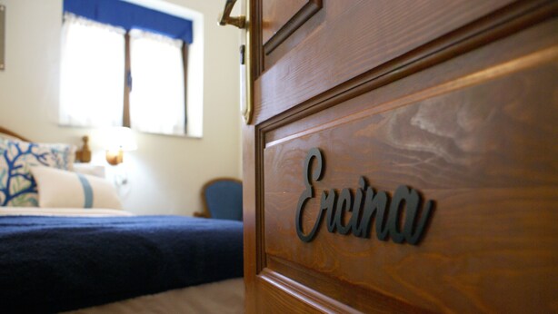 A wooden dresser with the name "Erin" carved on it, a bed with a blue blanket, and a window with blue curtains in the background. - Casa Rural Priena (Cangas de Onis)