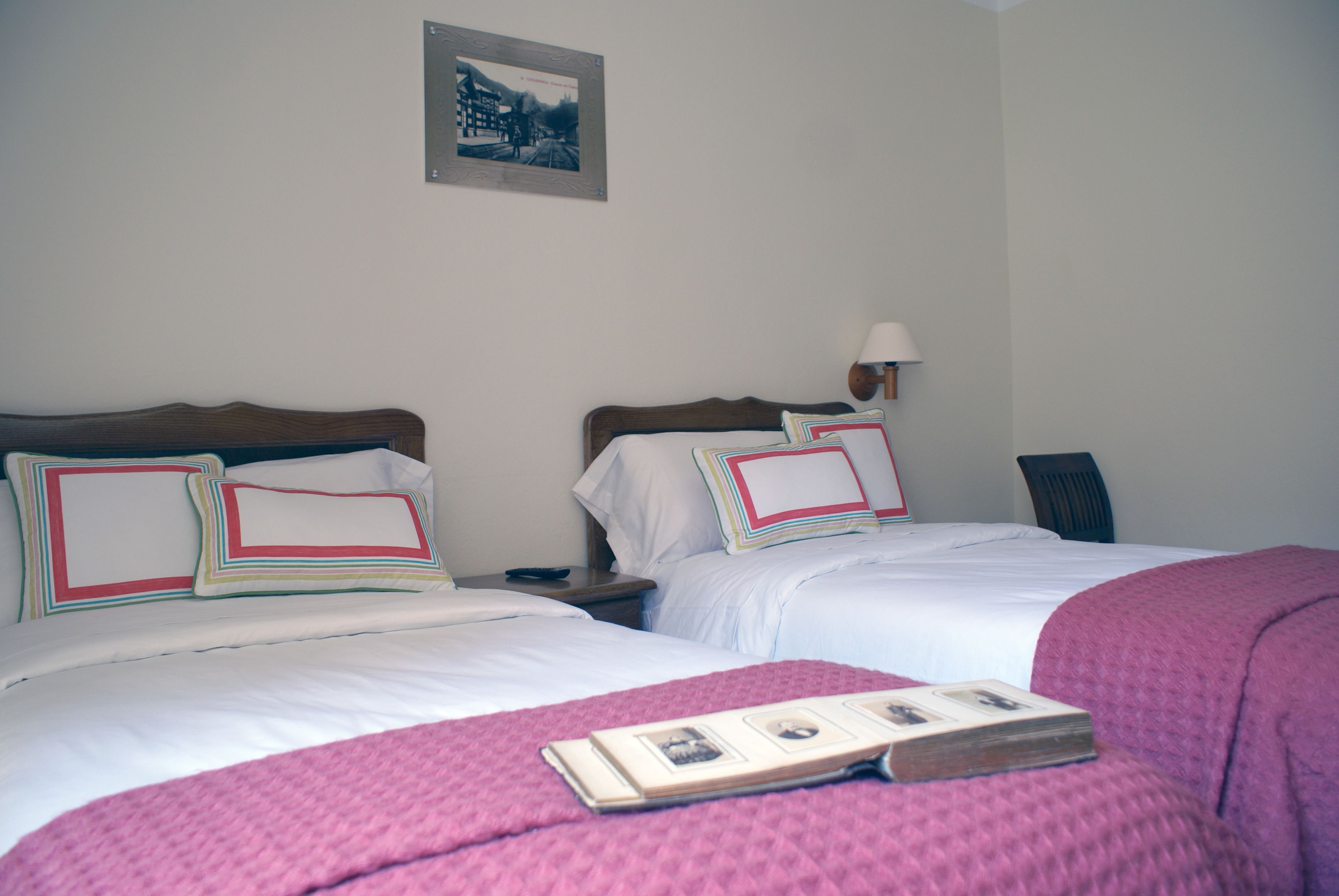 Une chambre d’hôtel avec deux lits, chacun doté d’une tête de lit en bois et de linge de lit blanc. On y trouve une table de chevet avec une lampe, un tableau encadré au mur et un livre sur le lit.