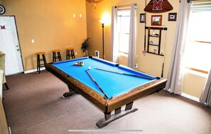 A pool table in a room with beige walls, a wooden floor, and a door with a 'M' sign.