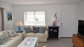 Ein Wohnzimmer mit einem beigefarbenen Sofa, einem weißen Couchtisch und einer TV-Konsole. Es gibt ein Fenster mit Jalousien, eine Lampe und eine Tür.