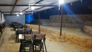 An indoor dining area with tables and chairs arranged in rows, a stone wall, and a stage area. - Edom camp (Dana)