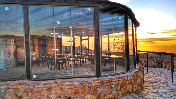 A glass-enclosed outdoor seating area with tables and chairs, stone walls, and a view of the sunset. - Edom camp (Dana)