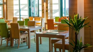 Un espace repas moderne avec des tables et des chaises en bois, de grandes fenêtres et une plante en pot.